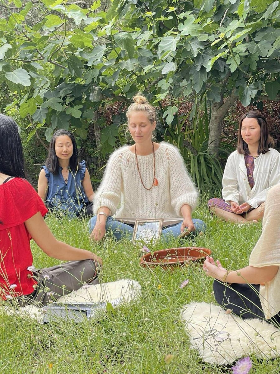 A group sitting together in the grass under a tree, Orama in the middle with a frame drum